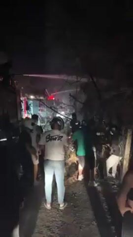 The video shows a nighttime scene with a crowd of people gathered around a fire truck. The truck is spraying water into a dark area, obscured by trees and branches. Most of the people are wearing casual clothing, and some are wearing hats or helmets. One person has a shirt with "SUPD" on it. A few individuals are wearing yellow and black shirts with the words "PAKET TAXI" on them. The overall atmosphere suggests an emergency situation.