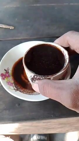 The video shows a close-up shot of a hand holding a white mug with a floral pattern and a dark liquid, likely coffee, inside. The mug is tilted over a white saucer, also with a floral pattern, and some of the liquid has spilled onto the saucer, forming a circular stain. The mug and saucer are placed on a dark wooden surface, possibly a table. The lighting suggests an outdoor or well-lit indoor environment. There are no other visible people or objects in the frame. The action is simple: the hand is holding and tilting the mug.