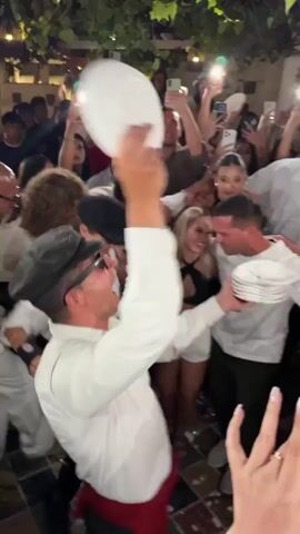 The video shows a group of people at what appears to be a celebration. In the foreground, a man wearing a white shirt, a black cap, and sunglasses is holding a white cake and appears to be interacting with another person in a white shirt. The cake is being held over a plate. Around them, many people are raising their phones to capture the moment, some with flash photography activated. The overall atmosphere seems festive and energetic, with people looking excited and engaged. The background suggests an indoor or semi-outdoor setting, possibly a restaurant or event space, with greenery and decorative elements visible. The people are dressed in casual to semi-formal attire, and the lighting indicates it is likely evening or a dimly lit venue.