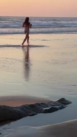 A young woman in a bikini jogs along the wet sand of a beach. The sun is setting, casting a warm glow on the scene and creating reflections on the water's surface. Waves roll in from the ocean in the background, where several other people can be seen in the water. The foreground is dominated by the sand, with a large rock formation partially submerged in a shallow pool of water. The overall mood is peaceful and serene.