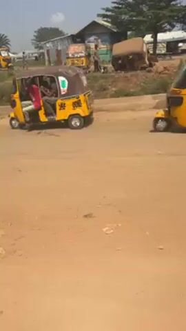 The video shows a dirt road lined with several yellow auto rickshaws.  One rickshaw has two occupants and is moving slowly. In the background, people are visible near simple buildings. A motorcycle drives past on the road.  The setting appears to be a rural area.