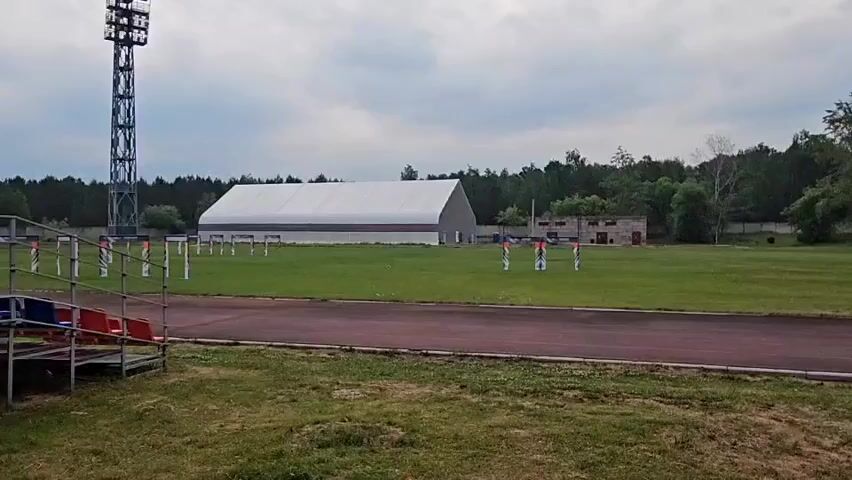 A wide shot captures a sports field under a cloudy sky. In the foreground, a section of red and blue stadium seating is visible. A red running track curves through the middle ground, bordered by a strip of grass. Beyond the track, the field is a expanse of green grass dotted with numerous white poles, each adorned with colorful flags. In the background, a large, white tent-like structure stands prominently, with a smaller, brick building to its right. A tall, metal tower, possibly a light tower, is positioned to the left of the frame. The field is surrounded by a dense line of trees.