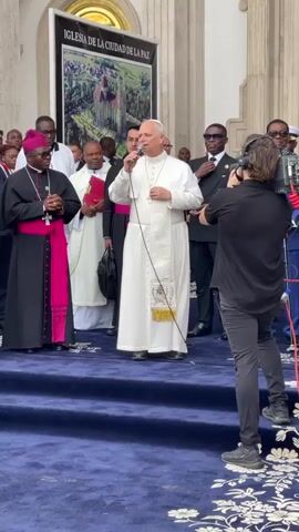 The image depicts a formal public event, likely religious or official, taking place outdoors or in a semi-enclosed grand space in Mongomo, Equatorial Guinea. The central figure, a man in white religious vestments including a white cassock, a short white cope, and an elaborately embroidered stole, is speaking into a microphone. He wears a pectoral cross.

To his left stands a man in black religious attire with a prominent pink sash or stole, also wearing a pectoral cross, with his hands clasped. Several other men, some in suits and others in religious garments, are visible in the background.

On the right, a cameraman, dressed in black, operates a professional video camera, capturing the event. The participants are standing on a platform covered with a blue carpet. In the background, a large white banner displays an aerial or architectural rendering of a building complex and the Spanish text "IGLESIA DE LA CIUDAD DE LA PAZ," which translates to "Church of the City of Peace." The architectural style visible in the background suggests a grand and formal building. The scene is illuminated by natural daylight.