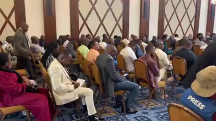 Here's a detailed description of the video:

**Scene:** The video shows a large room filled with people seated in chairs, presumably attending a conference or meeting. The room has an ornate design with patterned walls and carpeting.

**People:** The room is occupied by a diverse group of people, mostly men, dressed in a mix of casual and formal attire. Some are wearing suits, while others are in more relaxed clothing. Several attendees are taking notes. A man in a suit walks past the camera, appearing to be moving within the room. One individual in the foreground is wearing a blue vest with "PRESS" written on it.

**Objects:** The chairs are arranged in rows, and most are a tan color. Some individuals have papers or notebooks in their laps, indicating note-taking. The "PRESS" vest marks someone as a member of the media.

**Actions:** The primary action is people sitting and listening, suggesting a lecture or presentation is in progress. Some are actively taking notes, while others appear more relaxed. The man in the suit is walking through the room.

**Interactions:** The video mainly depicts individuals in a static listening posture. Interactions between attendees are not clearly visible.

**Environment:** The room's environment is formal and well-maintained. The walls have a patterned design, and the lighting seems adequate. Overall, the environment suggests an official gathering.