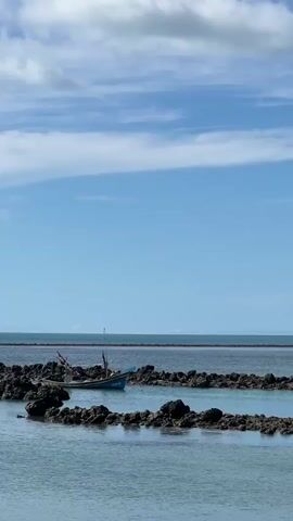 A small blue and white boat with two people aboard navigates through a calm, shallow body of water. The boat is surrounded by dark, rocky formations that create small channels and pools. The water is clear and reflects the blue sky, which is dotted with fluffy white clouds. In the distance, a thin strip of land is visible, and beyond that, the horizon meets the sky. The overall scene is peaceful and serene, with the boat appearing to be on a leisurely journey.
