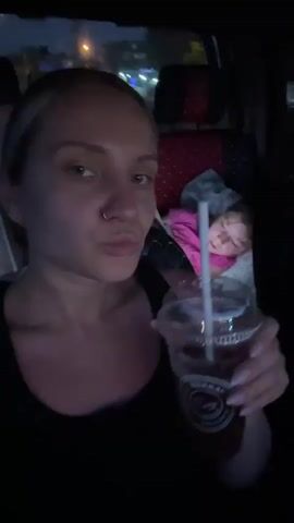In a dimly lit car interior, a woman with blonde hair and a nose ring sits in the front seat, taking a selfie video. She wears a black tank top and holds a clear plastic cup with a straw, taking a sip and then smiling at the camera. In the backseat, a young girl, wearing a pink shirt and glasses, appears to be asleep. Car seats and other items are visible in the background, suggesting a family car trip at night. Outside the car, streetlights are faintly visible.
