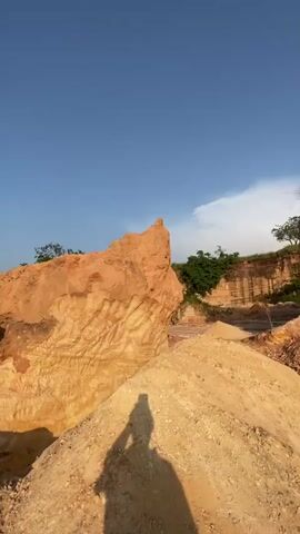 A person's shadow is cast on a pile of sand with a large rock formation in the background. The sky is blue with some white clouds. The rock formation has layers of orange and yellow. Trees are visible in the distance.