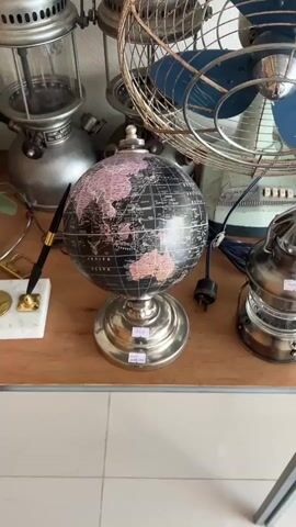 The video shows a hand reaching for a vintage globe that is on a wooden surface. The globe is black with pink land masses and white grid lines. There are price tags on the globe and a pen on a white marble base to the left. In the background, there are various vintage items including a fan, a lantern and what appears to be a lamp.
