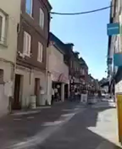 The video shows a street scene, possibly a town or city. It is daytime with a clear blue sky. Buildings line both sides of the street, appearing to be shops or businesses with storefronts visible. Some people are walking or standing on the sidewalk. There are streetlights and possibly some signs on the buildings. The camera appears to be moving, giving a sense of walking down the street.