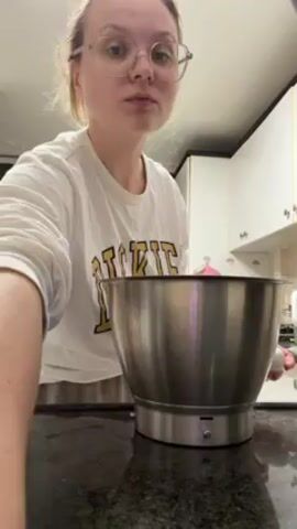 A young woman with glasses is in a kitchen, cracking eggs into a stainless steel mixing bowl on a black countertop. She wears a white long-sleeved shirt with "DICKIES" printed on it in gold and black. Behind her, white cabinets are visible, and there is a window with what appears to be a dark shade. A head of garlic sits on the counter to her left.