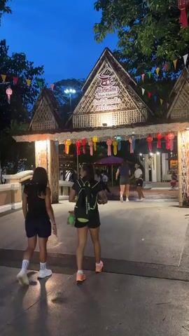 The video shows a group of young people walking through a decorative entrance at dusk. The entrance is made of bamboo and straw, with a triangular roof adorned with illuminated text in a foreign language. Colorful paper lanterns hang across the entryway, and small triangular flags are strung above. The ground is paved, and water reflects the lights, suggesting it may have recently rained or is wet from cleaning. Several individuals are walking away from the camera towards the entrance, dressed casually in shorts and t-shirts, some carrying backpacks. Trees with foliage are visible in the background, and the sky is a deep blue, indicating twilight. The overall atmosphere appears festive and welcoming.