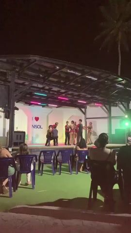 A group of dancers performs on a stage at night. The stage is lit by colorful lights. In the foreground, people sit in chairs watching the performance. The background shows a palm tree and the night sky. One dancer is in mid-air, having been lifted by another dancer. The dancers are wearing costumes. The stage has a backdrop with the text "I ❤️ INSOL".