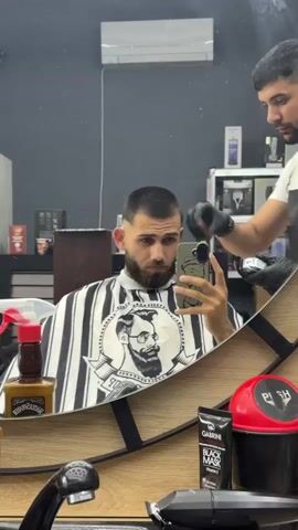The video captures a man getting a haircut in a barbershop, viewed through a mirror. The man, with a dark beard, is seated and wearing a barber cape featuring black and white stripes and a graphic of a bearded man with glasses. He holds a smartphone, seemingly recording or taking a selfie, while a barber works on his hair with electric clippers. The barber, wearing black gloves, stands behind him, focused on shaping his hair into a buzz cut or fade.

On the counter reflected in the mirror, several items are visible: a brown spray bottle, likely containing hair product, which resembles a repurposed "Jack Daniel's Tennessee Honey" bottle; a tube of "GABRINI BLACK MASK Vitamin E"; and a red container marked "PUSH." The mirror has a stylized wooden frame. In the background, an air conditioner is mounted on the wall, and shelves stocked with various barbershop products and equipment can be seen. The scene depicts a typical haircut appointment in Karadjovo, Bulgaria.