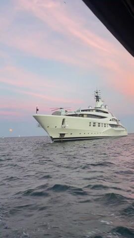 The video shows the view from a boat, passing by several large yachts on the ocean. The sky has a sunset with pink and purple clouds.  There is a hint of land in the background, with buildings and mountains. At least one of the yachts has a helicopter on the top deck.  The sea is slightly choppy.