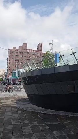 The video depicts a street scene in Taiwan. The camera is initially focused on a modern-looking, rounded structure with a dark gray, segmented exterior and a glass railing on top. The ground is paved with square tiles. The camera then pans to the left, revealing a busy intersection with many scooters parked along the sidewalk. The street is filled with people on scooters waiting for the traffic light to change. Buildings of varying heights and architectural styles line the streets, and street signs in both English and Chinese are visible. The sky is partly cloudy.