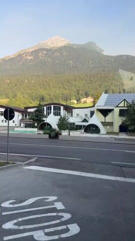 A vertical video shows a street scene with modern buildings in the foreground and a mountain range in the background. The street is paved and has white lane markings. A white vehicle is visible on the street. The buildings have unique architectural designs with curved white roofs. Trees and green spaces are interspersed between the buildings. The mountain range is covered in green vegetation at the base and rocky peaks at the top. The sky is clear and blue. The video pans slightly to the right, revealing more of the street and buildings.