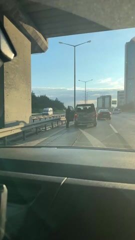 The video shows a view from inside a car on a highway. A person stands by a dark-colored van with damage to its rear. The van’s license plate is partially visible, reading “34 YT 199.” The person appears to be inspecting the damage. The highway has multiple lanes of traffic, with other cars passing by. A barrier separates the lanes from the side of the road. Overhead lighting is present. The sky is clear and the sun is shining.