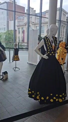 The video shows an indoor exhibition with several mannequins displaying outfits. The mannequins are arranged in a spacious area with large windows in the background, revealing a city skyline with buildings. The lighting appears to be natural daylight.

The first visible mannequin on the left is displaying a gold sequined jacket with heart-shaped embellishments and white pants. Behind it, another mannequin is wearing a black dress with yellow polka dots. A small mannequin in the center is also wearing a dress with a similar yellow and black polka dot pattern.

As the video pans, a mannequin in the foreground is adorned in a long, golden, shimmering dress with intricate detailing. Further back, a black mannequin is dressed in an avant-garde outfit featuring a short skirt made of coiled fabric and a spotted tail, all in yellow and black. Another mannequin on the right is wearing a flowing yellow dress with a train, also decorated with black polka dots.

The camera continues to pan, showcasing more mannequins. One mannequin is wearing a kimono-style orange dress with large black polka dots and a black sash. Another displays a black top with yellow sleeves and a yellow skirt with black polka dots. The video concludes by showing the back of a mannequin wearing a black dress with a train of yellow and black spotted fabric. Various display stands and barriers are also visible in the scene.