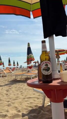 The video shows a beach scene with several rows of orange lounge chairs and closed umbrellas on a sandy beach. In the foreground, a bottle of Birra Moretti beer sits on a small red table. A person's foot is visible in the lower left corner of the frame, resting on what appears to be a towel. In the background, the ocean and a bright blue sky with scattered clouds are visible. Several people are scattered throughout the beach, some sitting on lounge chairs and others walking. The overall atmosphere is relaxed and suggests a summer vacation.