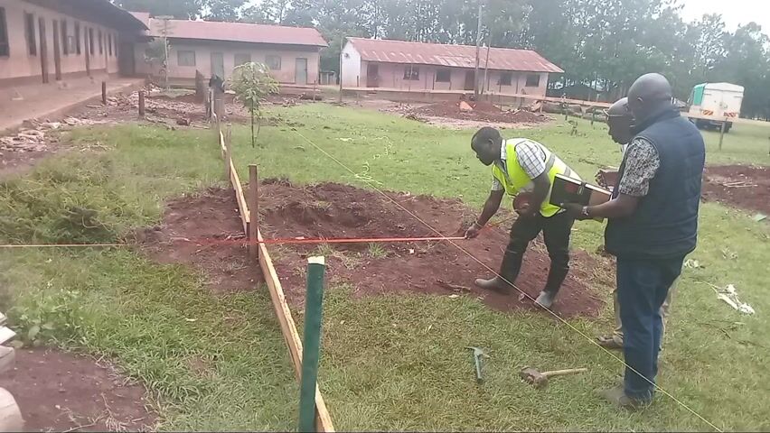 The video captures an outdoor scene featuring a group of men engaged in surveying or marking out an area on a grassy, partially dug-up plot. In the foreground, a man in a yellow safety vest and dark boots kneels and uses a yellow measuring tape along an orange string that demarcates a section of land. He then stands and points towards the distance. Another man in a light-colored shirt and khaki trousers holds papers, while a third man, wearing a patterned vest over a dark shirt and jeans, holds a notebook or tablet and gestures, seemingly directing or explaining something. Two other men stand nearby, observing the activity. Small hand tools, including a hammer and a pickaxe, are visible on the ground. In the background, several single-story buildings with corrugated metal roofs, typical of a school or community center, are visible. Trees line the distant horizon, and a white truck or trailer can be seen further back. The overall impression is that of a construction or development project in its initial stages. The sky appears overcast, suggesting either a cloudy day or early morning/late afternoon light. The ground has patches of exposed red-brown earth and green grass, with some wooden logs stacked on the left. The scene is set in Idakho South ward, Kenya.