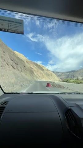 The video shows a car's interior dashboard and windshield view of a road trip through a mountainous landscape. The road curves to the left, with a steep, rocky mountain slope on the left and a riverbed on the right. The sky is blue with scattered white clouds. There are a few other vehicles on the road ahead and a small village or settlement visible in the distance. The timecode indicates the video starts at 00:00 and ends at 00:04.
