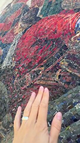 A close-up shot of a mosaic artwork is captured as a hand, adorned with a silver ring on the index finger, gently touches the surface. The mosaic appears to be made of small, irregularly shaped tiles in various shades of red, green, blue, and purple, creating a textured and visually dynamic pattern. The lighting suggests an indoor environment, possibly a museum or gallery, highlighting the intricate details and craftsmanship of the artwork.