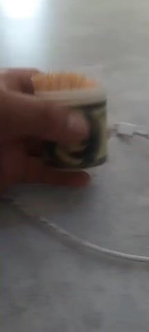 A hand holds a small container filled with toothpicks. The container is decorated with an image of Benjamin Franklin, resembling a hundred-dollar bill. A white cable with a USB-C connector is partially visible to the right of the container. The container is placed on a textured, light-colored surface, possibly a countertop or table. The video focuses on the container and the hand holding it, with a slight blur in the background.