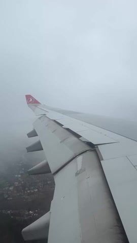 The video shows the wing of an airplane, specifically a Turkish Airlines plane, as it flies through a cloudy sky. Below, a town or city is visible through the mist. The plane's flaps are extended, suggesting it may be in the process of landing or taking off. The overall mood is overcast and grey due to the heavy cloud cover.