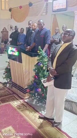 A man stands to the right of a podium holding a microphone with a sheet of paper in his left hand. The podium is decorated with Christmas trees with lights. Several people stand behind the podium. The man is wearing a brown suit jacket over a yellow shirt, white pants, and brown shoes. He is also wearing glasses. The podium has a sign on the front with multicolored lights. There is a red rug with gold trim in front of the podium. The floor appears to be made of marble. A ceiling fan is visible in the background. The event seems to be taking place inside a church.