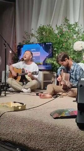 Two men are seated on a raised, carpeted platform, playing acoustic guitars. The man on the left is singing into a microphone, while the man on the right is focused on his guitar. Behind them, a large television screen displays a scenic image. The background is decorated with lush greenery and white flowers, with draped white fabric. The overall setting appears to be an indoor performance space.
