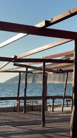 The video features a slow, horizontal pan across an empty, open-air wooden structure on a coastline. The foreground is dominated by a weathered wooden deck floor, with sunlight casting long shadows from the structure's vertical posts and horizontal beams. This wooden framework, possibly a pergola or a dismantled beach bar, offers an unobstructed view of the sea. Beyond the structure, the calm, blue expanse of the sea stretches out to a distant rugged coastline with cliffs or mountains. The sky is clear and bright blue, suggesting fair weather. The scene appears serene and peaceful, without any people present. The setting is Bratica - Braticë, Montenegro.
