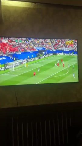 The video shows a large television screen displaying a live soccer match. The game is being played on a green grass field with white lines marking the boundaries and penalty areas. Two teams are playing; one team is wearing red uniforms, and the other team is wearing white uniforms. Several players are visible on the field, actively engaged in the game. A player in a red uniform is near the goal on the left side of the screen, potentially in the process of kicking the ball. A goalkeeper in a yellow uniform is in the net. The score displayed on the screen indicates 1-0. The background of the stadium shows spectators in red seats. The television screen itself is large and mounted on a wall with a textured wallpaper pattern. Below the TV, a dark wooden headboard of a bed is visible, with vertical slats.
