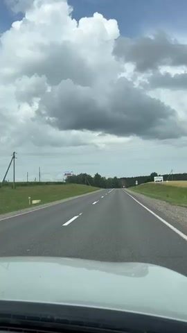 The video shows a view from inside a moving car on a paved road. The road has white lane markings and a speed limit sign of 50 is visible on the right side. There are fields of green grass and crops on both sides of the road, with trees and a cloudy sky in the distance. A billboard is visible on the left side of the road. The car's dashboard is visible in the foreground.