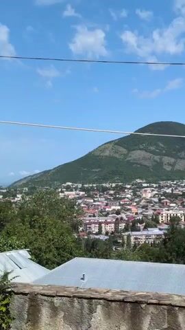 This video captures a panoramic view of a town nestled at the foot of a lush green mountain under a clear blue sky with scattered white clouds. The time of day appears to be late morning or early afternoon, judging by the bright sunlight. The town is characterized by a dense collection of buildings with red-tiled roofs, interspersed with trees. Some larger, more modern buildings with white facades and multiple stories are visible, along with what appears to be a tower or minaret, hinting at a historical or religious significance. In the foreground, a rustic stone wall and a section of a corrugated metal roof provide a grounding element, suggesting the viewpoint is from a slightly elevated position, possibly a balcony or a hillside. The overall impression is one of a peaceful, scenic town with a backdrop of imposing natural beauty. The weather seems pleasant and sunny, contributing to a calm and inviting atmosphere. There is no discernible text in the video.
