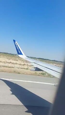 The video shows the wing of an airplane on a tarmac. The plane has the logo "flypgs.com" on its tail fin. The sky is clear and blue. The ground is dry and sandy with some sparse vegetation. A shadow of the airplane wing is cast on the tarmac. The plane is likely in the process of takeoff or landing.
