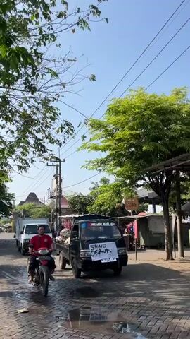 A motorcycle is seen from the side moving forward on a paved road. The rider is wearing a red shirt and dark shorts. Behind the motorcycle, a black pickup truck with a white sign that reads "SANO JERUK" is visible. Further down the road, a white delivery truck is also moving forward. The scene appears to be outdoors on a sunny day with trees and buildings lining the street. Electrical wires are strung across the sky.