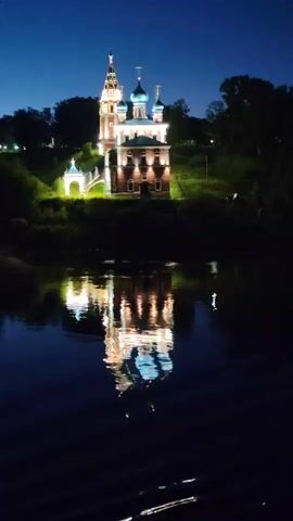 This is a nighttime video of the Resurrection Cathedral in Tutaev, Russia. The cathedral is illuminated, highlighting its intricate architecture with a tall, ornate bell tower and multiple domes. The building sits atop a hill, with lush greenery surrounding it. In the foreground, a dark body of water reflects the illuminated cathedral, creating a stunning mirror image. The sky is a deep, clear blue, indicative of a twilight or night scene. The overall mood is serene and majestic, with the lights of the cathedral creating a warm glow against the dark landscape. There is no visible text in the video.