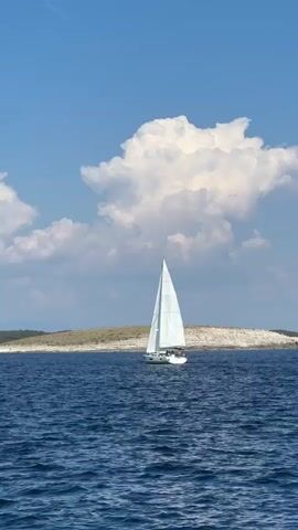 A sailboat with a white sail moves through blue water under a sky with scattered white clouds. An island with sparse vegetation is visible in the distance. The camera is angled slightly upward and is relatively static.