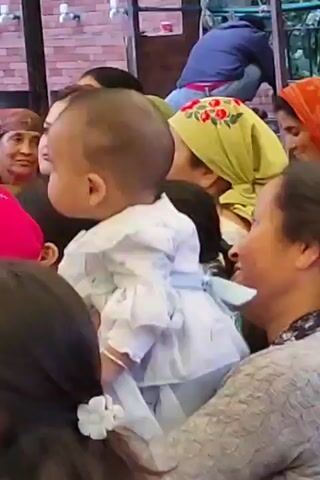 The video shows a baby held by an adult in a crowded outdoor setting. The baby is wearing a white dress. Several people, mostly women, are visible in the background, some wearing headscarves. The environment appears to be an event or gathering, possibly a celebration. The people are looking towards the baby and interacting with each other.
