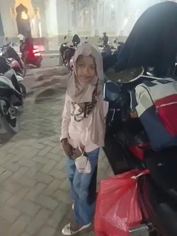 A young girl in a light pink hijab and light blue jeans stands in front of a parked motorcycle, holding a white pouch in her right hand. Behind her, a collection of motorcycles are parked in a lot adjacent to a mosque. The mosque is illuminated by artificial lights, and the sky is dark, indicating it is nighttime. The ground is paved with light-colored bricks. Red plastic bags are visible on the ground near the motorcycles.