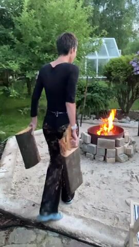 A person wearing a black long-sleeved shirt and black lace pants with a white watch on their left wrist is standing in a backyard, holding two pieces of firewood. The person is facing away from the camera and is in the process of adding the firewood to a lit fire pit. The fire pit is made of metal and is surrounded by bricks. There are trees and bushes in the background, as well as a greenhouse. The ground is a concrete patio. The person places the first piece of wood into the fire pit, and then bends down to pick up the second piece, which they also add to the fire.