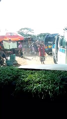 The video shows a street scene in an African country. The view is from inside a vehicle, through the windshield. A row of greenery is visible at the bottom of the frame, obscuring the lower part of the scene. In the foreground, on the left side, there is a makeshift stall with a red and white umbrella. People are sitting around it. Further back, there are more stalls and people milling about. A woman in a red top walks towards the camera, carrying a plastic bag with what appears to be coconuts in it. Parked to the right, there are vans or minibuses, one with the word "PRESTIGE" visible. The ground is dusty. The sky is bright.