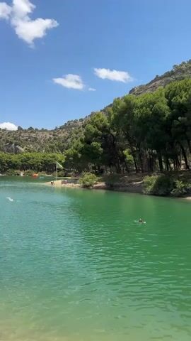 A vertical, full shot of a lake with clear green water under a blue sky with white clouds. The lake is surrounded by a lush green forest and a rocky, treed hillside. In the distance, several people are swimming and wading in the lake. Some are near a small beach with umbrellas and what appears to be inflatable water toys. In the foreground, two people are sitting on the edge of the water. One person is standing, facing away from the camera, and looking out at the lake. The other person is sitting with their legs in the water, also facing away. The sun is shining brightly, creating reflections on the water.
