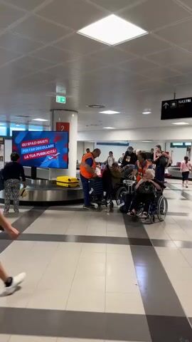The video shows an airport baggage claim area. Multiple people in wheelchairs, attended by airport staff wearing orange vests, are gathered near a baggage carousel. Some passengers are retrieving their luggage. There's a large digital screen on a column displaying an advertisement. People are moving around the area, including a young man walking in the foreground. Signs indicating "Belt" and an exit are visible. The lighting is bright and the floor is tiled.
