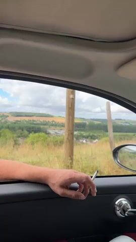 The video shows a person's arm and hand resting on the inside of a car door, with a cigarette held between their fingers. The view through the open car window reveals a rural landscape with rolling hills, fields of crops, trees, and a village in the distance. The sky is partly cloudy, and a wooden post is visible in the foreground outside the window. The hand moves slightly as the video progresses.