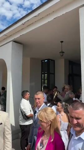 A group of people are gathered outside a building. A man in a white shirt is playing a saxophone. A man in a light-colored suit is walking towards the camera. A woman in a pink jacket is standing next to him. Another woman in a white dress is in the background. The weather is sunny with some clouds in the sky.