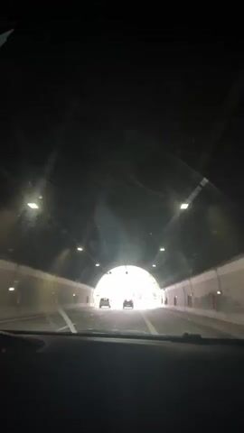 The video shows a point-of-view shot from inside a car driving through a tunnel and then onto a highway. Initially, the car is in a dark tunnel with artificial lighting. Two cars are visible ahead, heading towards the tunnel exit. As the car emerges from the tunnel, daylight appears, revealing a green, mountainous landscape. The car then joins a highway with multiple lanes. More cars are seen on the highway, and there are streetlights lining the road. The sky is overcast with clouds. The windshield has some smudges and reflections.