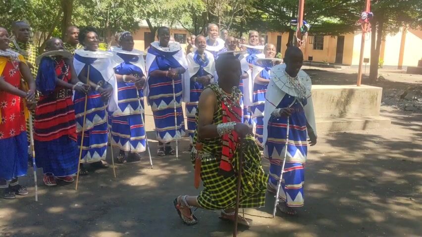 A group of about 15 people are assembled outside, possibly in a schoolyard.  The majority of the group are dressed similarly in blue and white skirts, white shawls, and large white beaded neckpieces. They are holding walking sticks.  One person stands out; dressed in a yellow and black checkered garment and decorated with what appears to be a silver arm cuff. He kneels before one of the women, who reciprocates the gesture. All seem to be participating in a ceremony or performance.  The area around them is shaded by trees, and a building is visible in the background. The atmosphere appears joyful, with some individuals smiling.