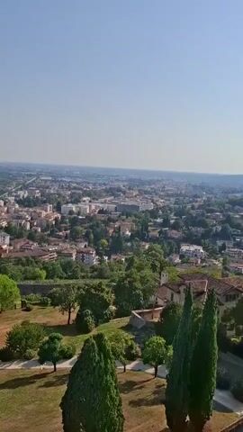 The video shows an aerial view of a town with buildings and trees. The sky is clear and blue. There are no people or animals visible. The camera is panning slightly to the right, giving a sweeping view of the landscape. The buildings are mostly made of brick and have red tiled roofs. There are a few tall cypress trees in the foreground, and a number of green trees scattered throughout the scene. The ground in the foreground is covered with dry grass and some dirt.