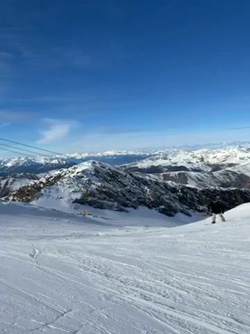 The video shows a skier skiing down a snow-covered mountain on a sunny day. In the background are snow-covered mountain peaks and a blue sky with some clouds. Ski lift cables are visible in the upper left.